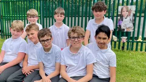 BBC Five boys in white polo shirts sit on a bench and another three stand behind them. Behind the group is a green metal fence with a large poster of a young boy and teddies attached to it.