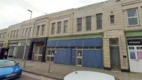 The tiled front of the Art Deco building comprises two floors, with shops on the ground floor and multiple windows on the first floor.
