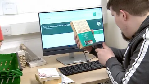 Chris is pictured using a handheld barcode scanner to scan the back of a paperback book at a desk. There is a computer screen in front of him.