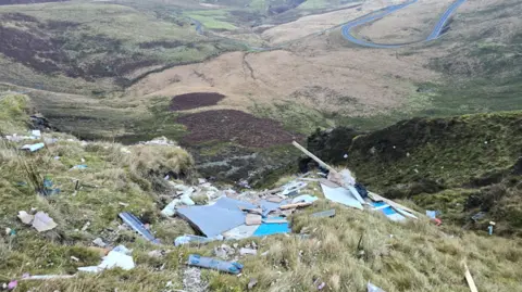 Nathan Dixon A view of the dumped waste on the mountain looking down on the valley below. A winding road can be seen in the distance. 