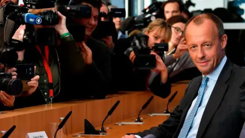 Getty Images Merz sits in a blue suit and tie, staring at the camera, while TV cameras are designed from behind