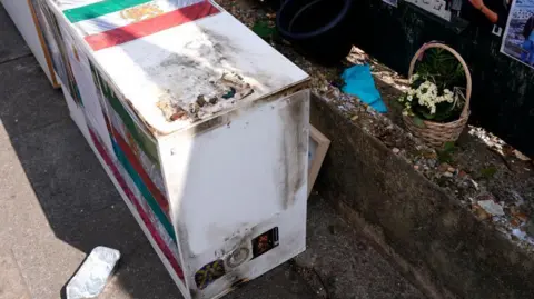 PA A cabinet with images of the Iranian flag attached is covered in dark charred markings. Flowers can be seen lining a small ledge.