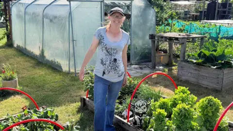 A woman in a grey top and blue jeans stands in the allotment. Behind her is a green house and some wooden planters. Either side of her are wooden planters with red hoops over the top. A small highland cow picture is in the planter near her leg.