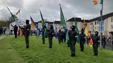 A group of people are dressed in black jackets, black berets, dark glasses, face coverings and khaki combat trousers. They are holding flags and standing on a grassy area. The masked men are being watched by a crowd of people in front a housing estate.  