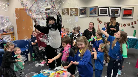 A group of children playing with toys as the Library cat blows bubbles around them. Most of the children are in school uniform which is blue jumpers, yellow tops and green tops. The children are laughing and catching the bubbles. 