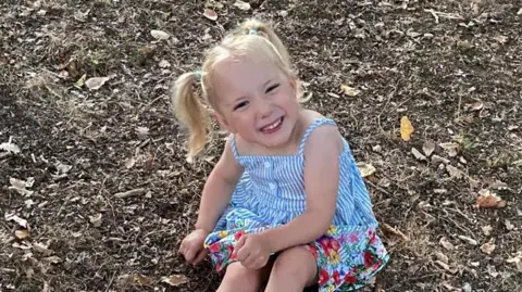 Bella smiles and looks up at the camera while she sits on the floor. She has blonde hair which has been tied up in pigtails. She is wearing a blue and white striped summer dress. 