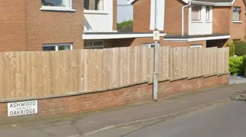 An unpainted fence runs above a knee-height brick wall, leading into a housing estate. Behind the fence and wall are a few houses.