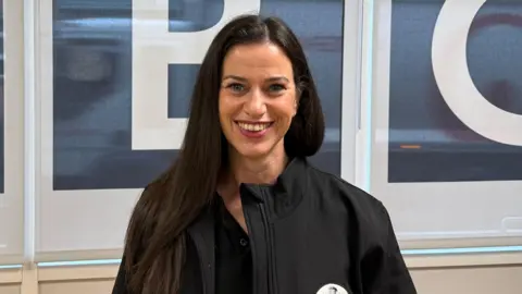 BBC/Elliott Green A woman with long dark hair and a black fleece jacket smiles at the camera. She has lipstick on. Behind her are some blinds with the BBC branding.