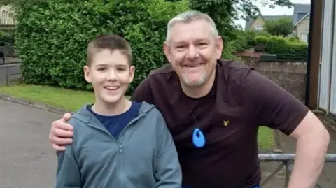 Family photograph Teenager, Dexter, (left) wearing a grey coat and blue pants. He has brown hair. He is being hugged by John Davidson with grey hair. He is wearing a maroon t-shirt, around his neck is a blue silicone toy. They are stood in front of some trees. They are both smiling.
