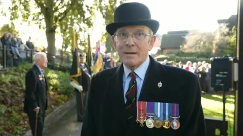 BBC Lt Col Mark Jackson stood wearing a black bowler hat and black coat, with four army medals on display 