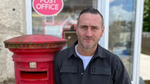 BBC Will Mellor stood outside a Post Office leaning on a post box