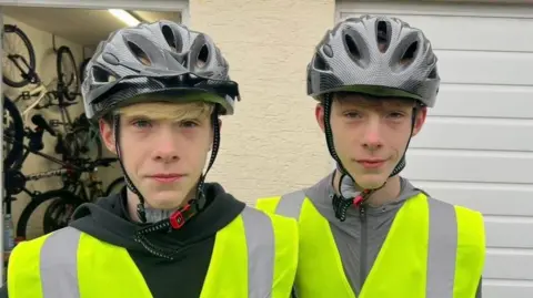 Twins Thomas and Travis looking into the camera. They are both wearing grey bike helmets and neon green hi-vis jackets. Travis, who is on the left, is wearing a black hoodie and Thomas is wearing a grey zipper. 
