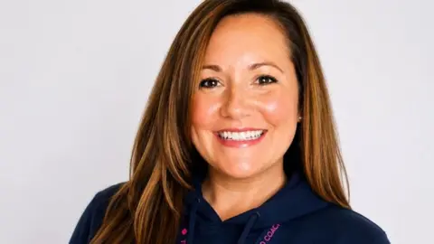 Head and shoulders of Sam Wright. She is smiling and has long straight brown hair and is wearing a dark blue zip up top