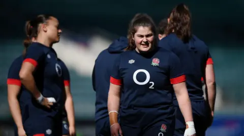 Rugby Football Union Maud Muir during a game. Other players are also seen behind her. She is looking away from the camera. It's a sunny day.