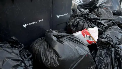 Getty Images Black bin bags are stacked high around a dark wheelie bin bearing the Birmingham City Council logo and heart emblem. A cardboard box pokes out from among the bags, with other packaging visible in the pile. 