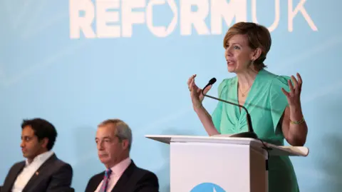 Andrea Jenkyns, wearing a green dress, speaks at a podium. On her right are sitting Nigel Farage, wearing a suit and pink shirt, and Zia Yusuf, wearing a suit and white shirt