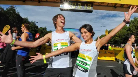 Jon Egging Trust Dr Emma Egging and Professor Brian Cox at the finish line of a marathon race. Both are smiling with their hands stretched out and wearing white running vests, with a "Jon Egging Trust" logo and a race number.