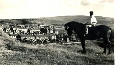 BBC A black and white photo of a man sitting on a horse overlooking Merthyr Tydfil, with hills in the distance.