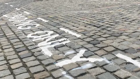 The words Loading Only have been painted onto cobbles but the paint is smudged in many places. Three benches are at the top of the picture 
