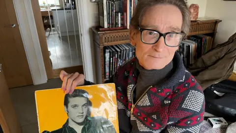 Giles Moriarty, 57 is wearing glasses and sitting in a chair holding an Elvis Presley album showing the young Elvis in black and white on a yellow background. Giles has brown hair and a dark rolled neck sweater with a red black and white patterned cardigan over the top, there is a dark wooden book case behind him filled with Elvis books.