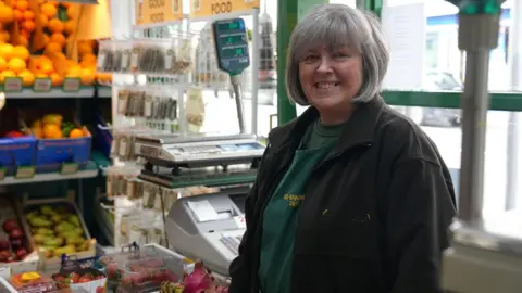BBC Hayley Morgan is standing behind her counter in her greengrocers. She has a grey bob and is wearing green uniform with a black jacket over the top. 