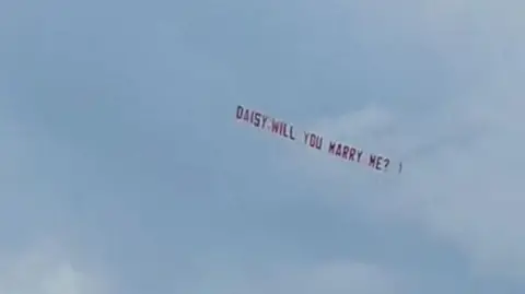 A sky banner being trailed by a plane reads 'Daisy will you marry me?' on a blue sky backdrop