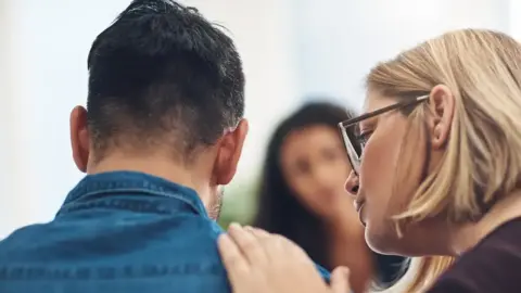 A man in a blue shirt with his back to the camera is comforted by a woman with short blonde hair wearing glasses. She is patting his right shoulder. A third person, a woman, is in the background but is blurry.
