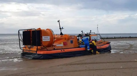 Caroline Robb An RNLI hovercraft on a wet, flat beach with crew attending to people inside. The sea looks calm in the background.