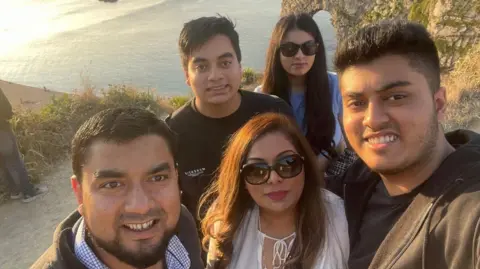 Hafsa Begum A 45-year-old woman is standing next to her husband and three children on a coastal path with the sea in the background. The sun is shining and they are all smiling. There is a sea arch made by erosion in the background.