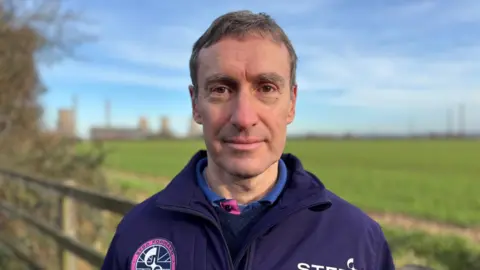 CEO of UKIFS Paul Methven is pictured wearing a company-embroidered fleece top. He has short brown hair. In the background, blurred, are green fields and the power station.