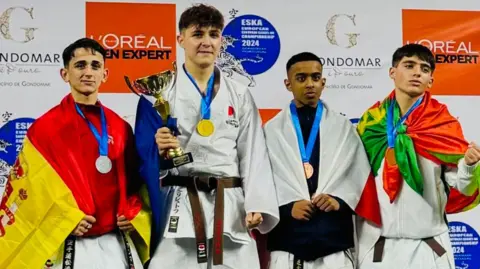 Four boys are stood on a podium at the European Shotokan Karate-do Association European Championships. Medals are hanging from their necks, and draped around their shoulders are their national flags. The winner is also holding a trophy. Mayon has England's flag wrapped around him, and looks proud with the bronze medal.