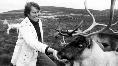 Tony Soper - a black and white image of a man with ear length long brown hair, smiling whilst feeding a reindeer. He is wearing a black turtle neck top with a white overcoat, in a field with reindeers.