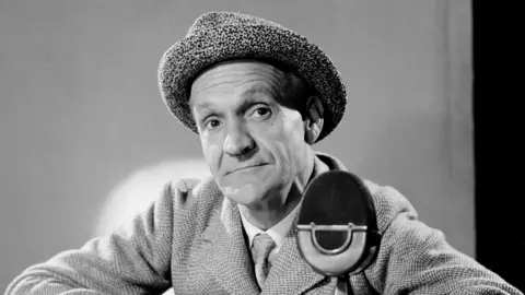 A black and white image of Stanley Unwin, who is sitting by a microphone. He is leaning on a table and wearing a suit and hat.