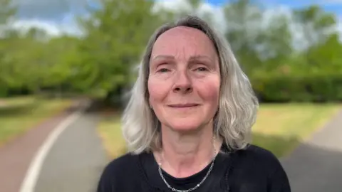 Head and shoulders image of Sharon Lewis. She has shoulder-length white hair and is wearing a black top and silver necklace.
