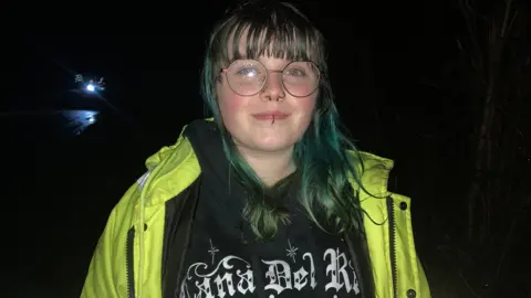 Maisie is standing in the dark wearing a Lana Del Ray shirt and high-vis jacket. She has long, dyed hair.