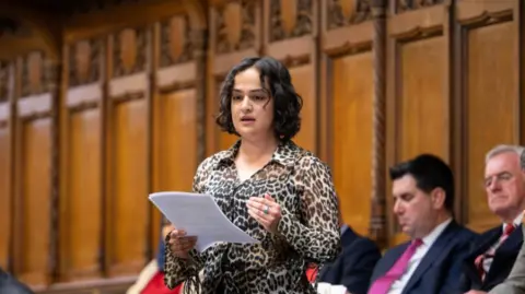 UK Parliament Whittome is a young woman with bobbed, curly black hair and wears a leopard print shirt.