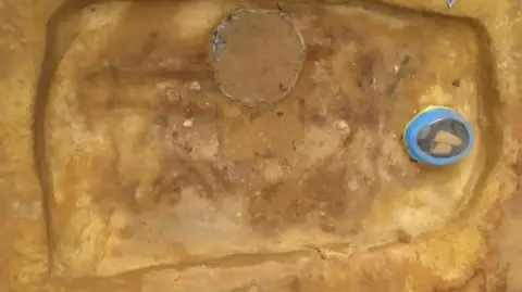 OCA A side view of two sand skeletons emerging from a burial chamber, excavated from sandy soil, with the head end to the right. The faint outlines of a person lying on their back is at that top. Across its torso and upper legs the edge of a large copper vessel is emerging. Beneath is a person lying on their side, with their legs bent. 