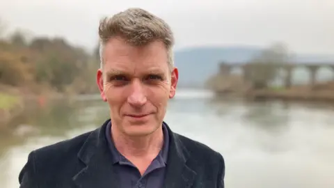 Wye and USK Foundation CEO Simon Evans looks at a black jacket and a camera in front of the Wye River in Monmouth in Purple Shirt.