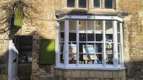 Tarot Crick Stone-fronted building with a bay window displaying artwork; a green hanging sign reads ‘Jackdaw Gallery’ above the shop window on a sunny street.