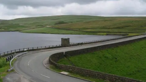 Anderson/Geograph Huddersfield Road snakes above the Dowry reservoir dam, with a basin of water seen behind the route, with green hills in the distances on an overcast day.