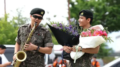 BTS members RM and V greet fans with salutes and a saxophone performance