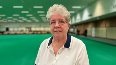 A woman with grey hair and a white shirt in front of a bowling green.