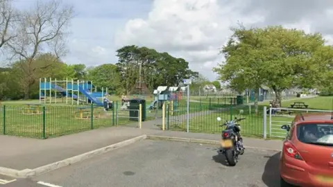 Google Street View image of Hayle Recreational Ground in Cornwall. A play park is in the background. A black motorbike and dark orange car are parked up outside the park's grounds. 