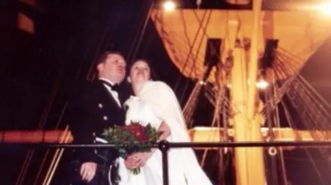 Family Chris Egan wearing a dark suit with his wife, wearing a wedding dress on a boat. Chris has short dark hair and wears what looks like the tartan of a kilt. Behind them, the mast of the ship is lit up yellow by lights.