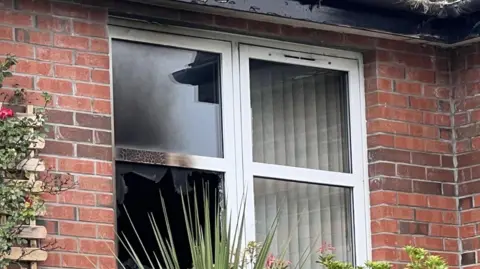 BBC Damaged window, inside white frame, at red brick bungalow in the Forthriver Road area of Belfast.
