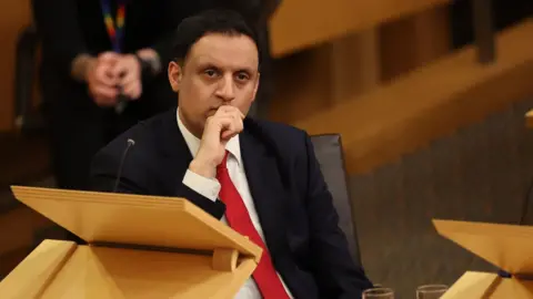 PA Media Anas Sarwar sits behind a desk in the Scottish Parliament chamber. His right hand is over his mouth and he is wearing a black suit, white shirt and red tie.