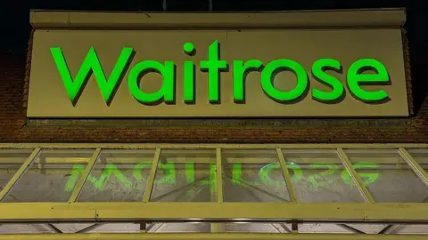 A bright green Waitrose sign on the roof of a shopping centre.