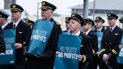 Airline pilots wearing a black uniform with a hat and gold embellishments hold blue signs saying 'No pilots no profits'