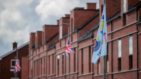 The PA media sits in front of a line of three lamppost red brick houses. There are three flags on these lampposts. There are two identical, they are red, white and blue union jacks. The second is a mild blue UDA flag, with a red hand of the olster at its center. The flags partially flutter into the air. 