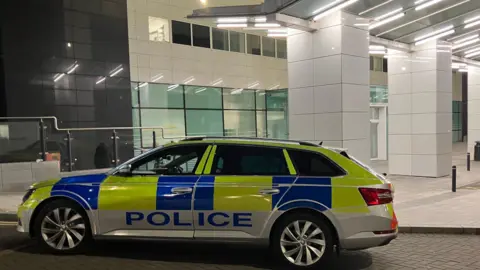 A police car parked outside the Royal Victoria Hospital Emergency Department. The building is white. The police car is yellow and blue and says POLICE along the side of it.